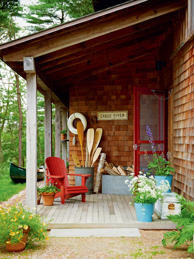 Wooden porch with paddles, firewood, potted flowers, a red chair, and a canoe in the background.