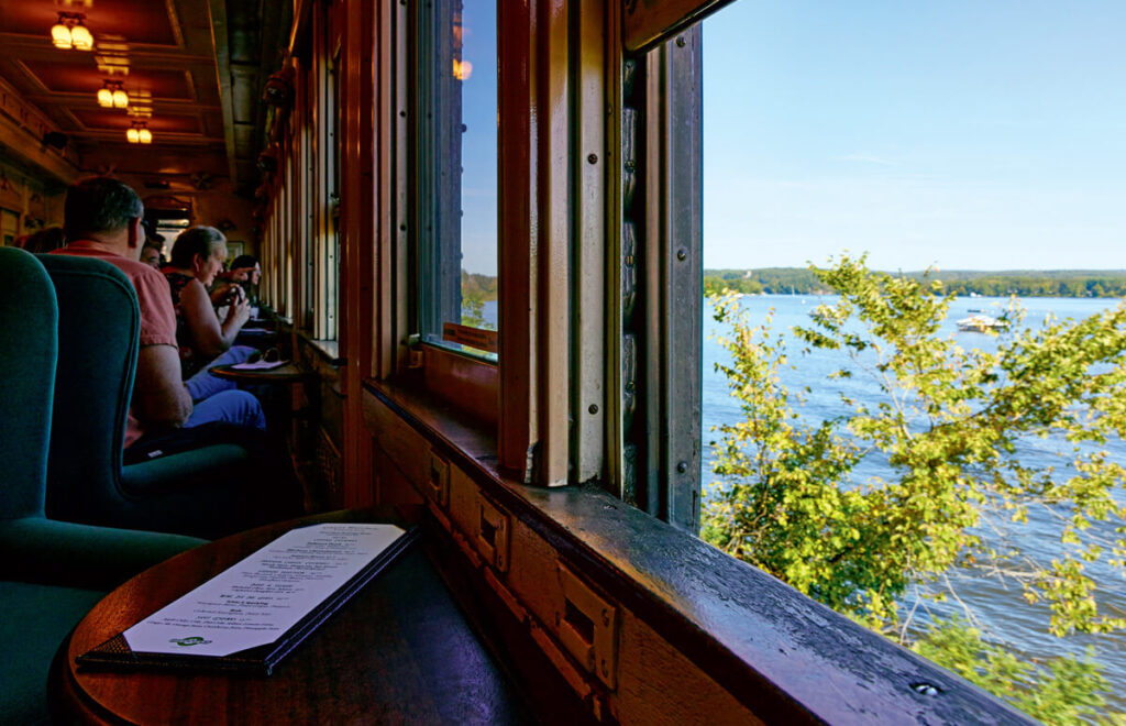 All Aboard the Essex Steam Train & Riverboat in Essex, Connecticut. People dining inside a train car with large windows overlooking a scenic river and trees.