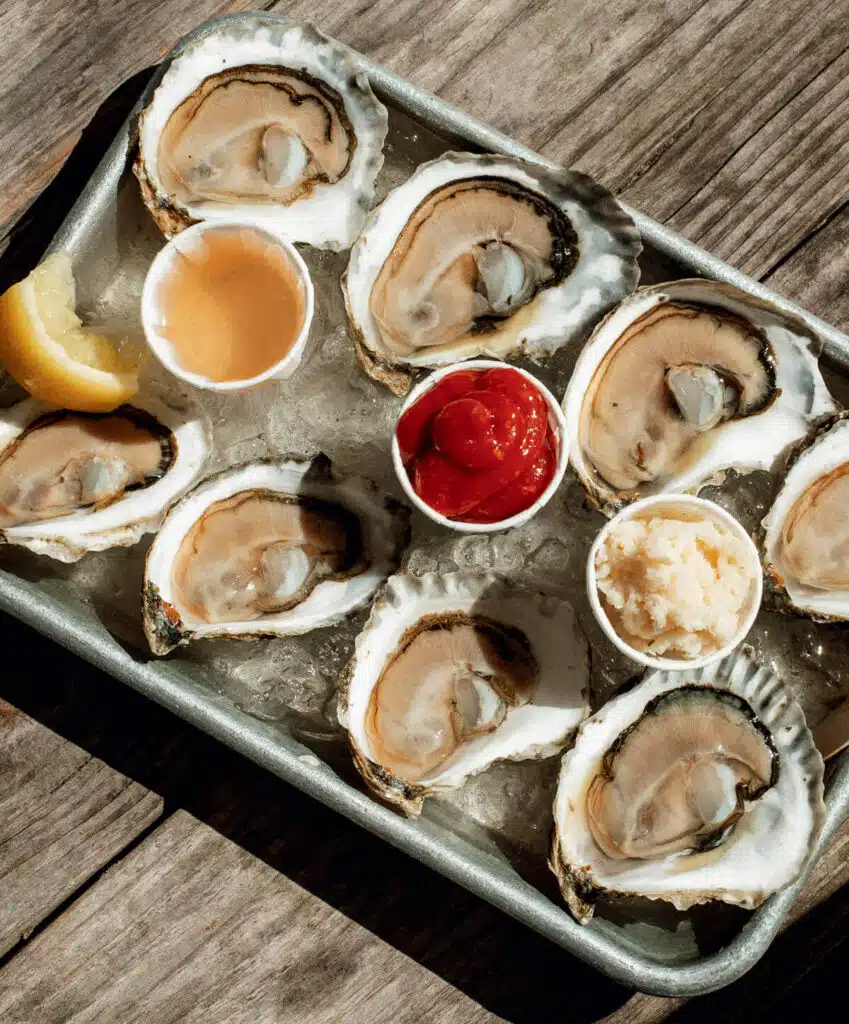 A tray of fresh oysters on ice with lemon, cocktail sauce, vinegar, and horseradish, on a wooden table.