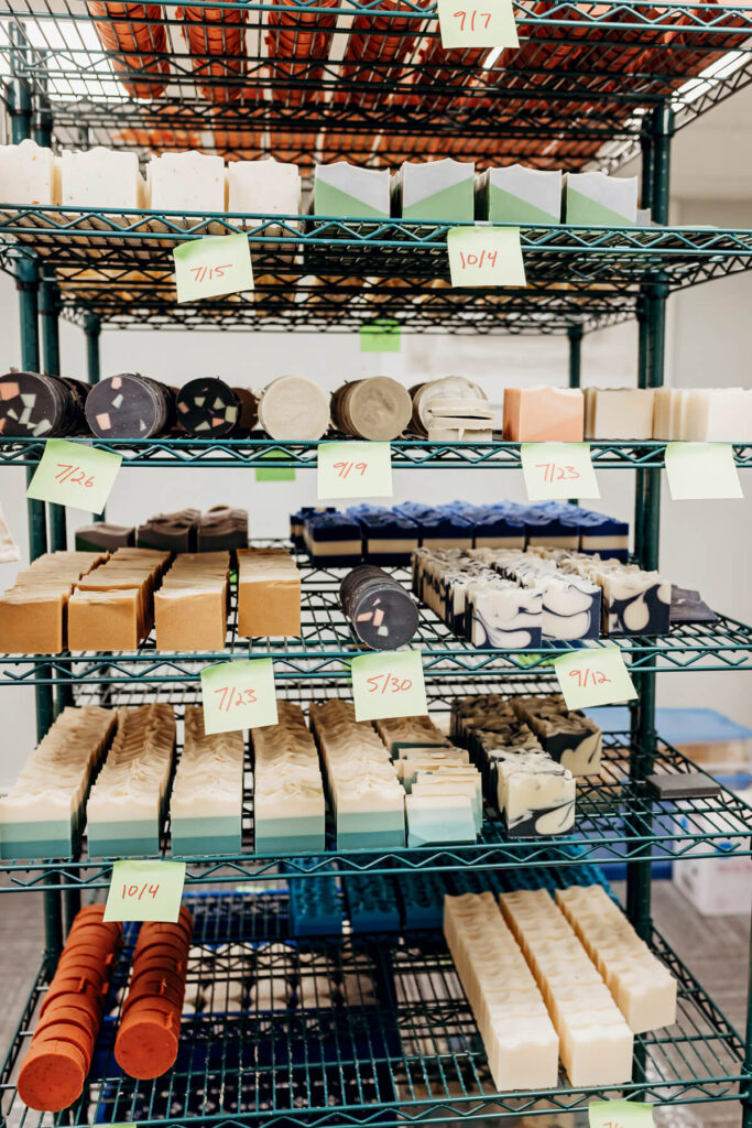 Shelves filled with handmade soaps in various shapes and colors, labeled with dates on sticky notes.