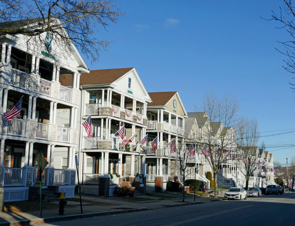 Row of houses with American flags displayed on porches along a sunny, tree-lined residential street.