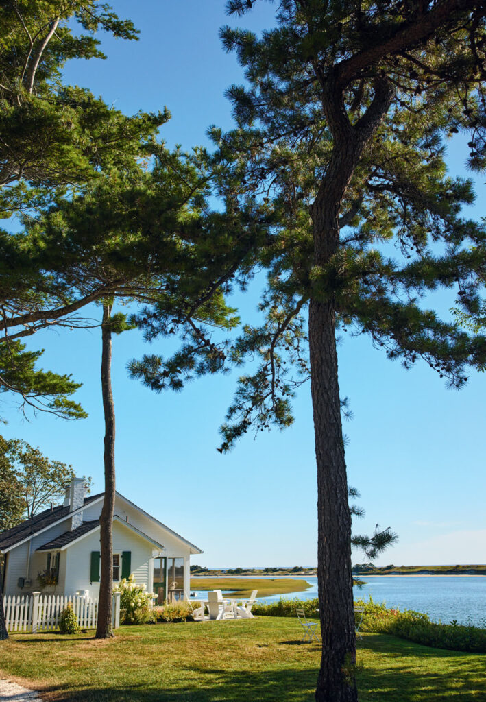White cottage with green shutters by a calm lake, surrounded by trees and outdoor chairs on a sunny day.