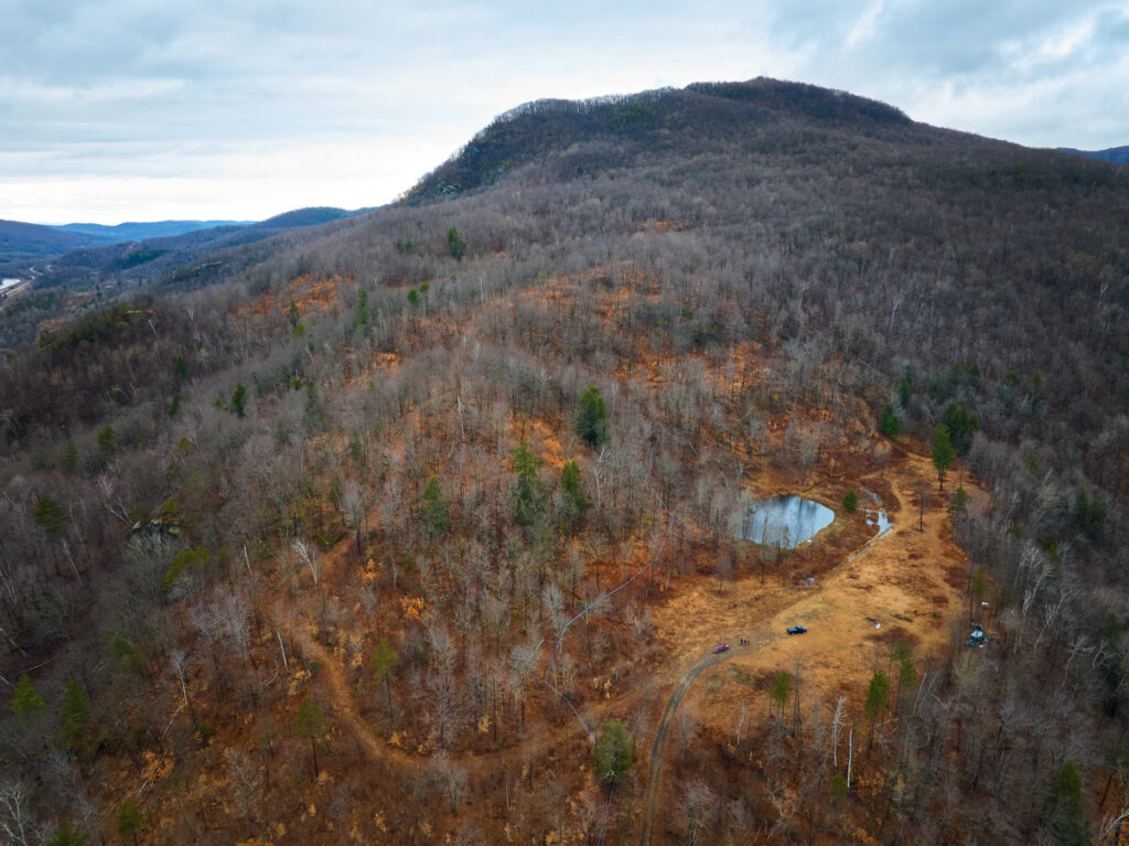 Aerial view of a hill with bare trees, a small pond, and a few vehicles on a cloudy day.