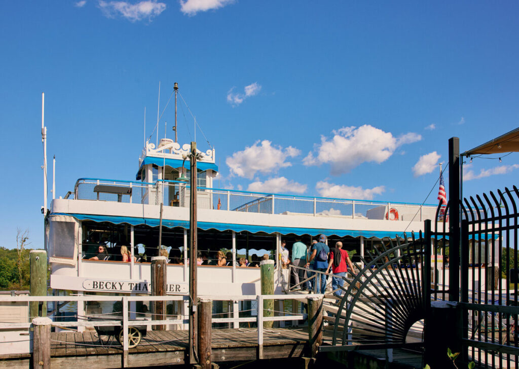 People boarding a white and blue riverboat named "Becky Thatcher" at a dock under a clear blue sky.