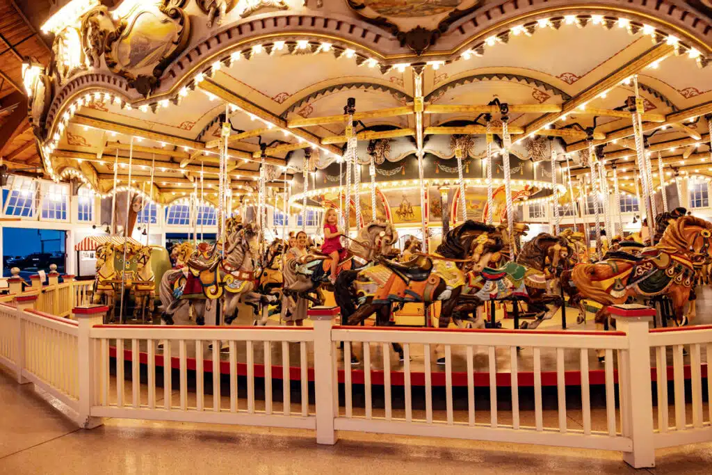 A brightly lit, colorful indoor carousel with ornate horses, surrounded by a white fence.