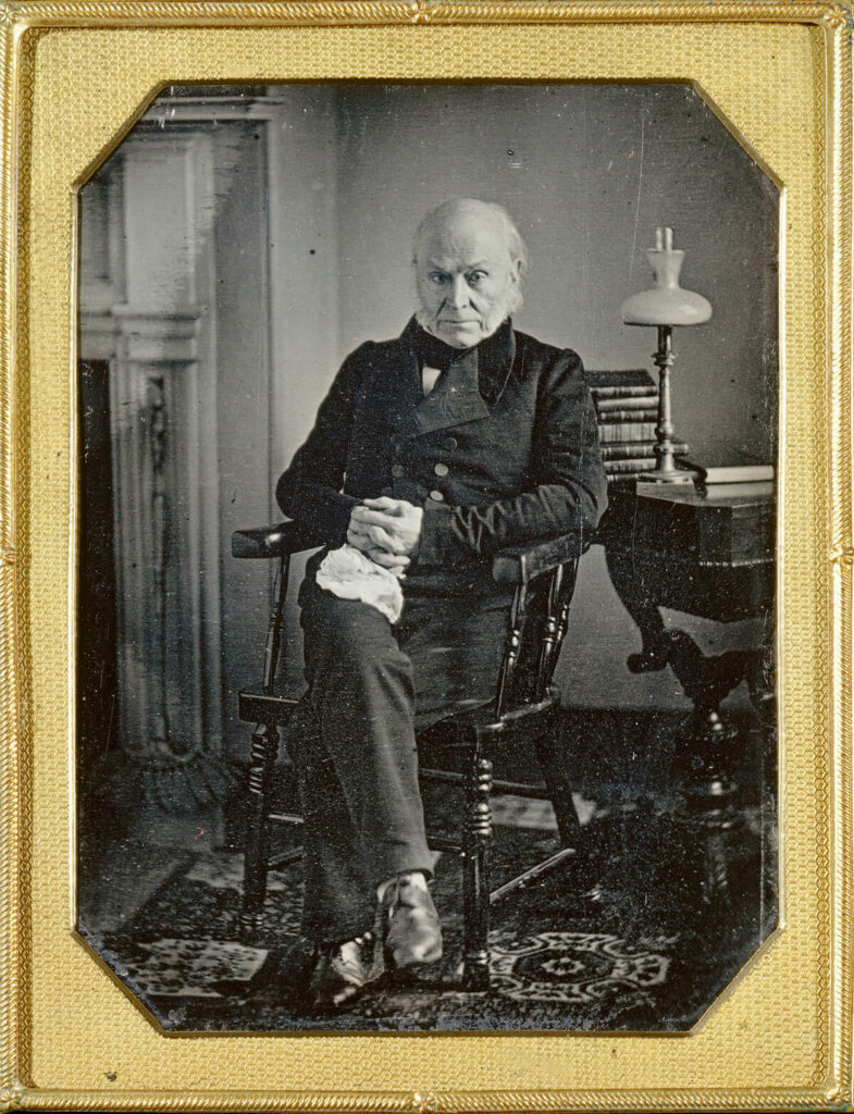Elderly man in formal 19th-century attire sits on a chair beside a table with books and a lamp.