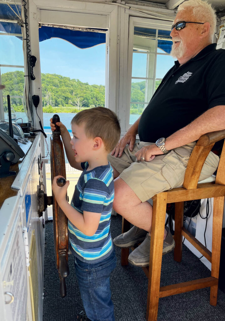 A young boy steers a boat while an older man watches, both inside the boat’s cabin on a sunny day.