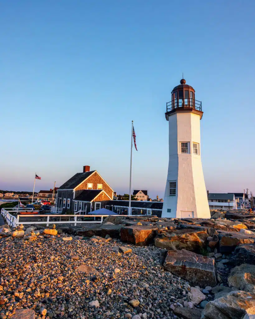 Tall white lighthouse and rustic building by rocky shore at sunset, with flagpole and clear blue sky.