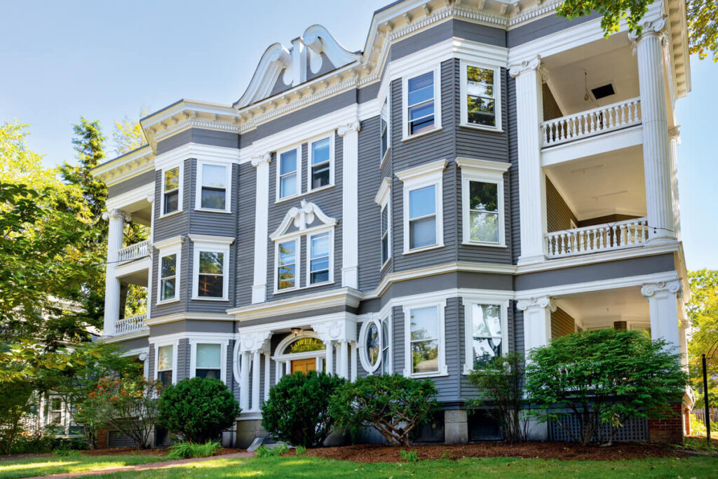 Gray three-story historic apartment building with white trim, large windows, and columns surrounded by greenery.