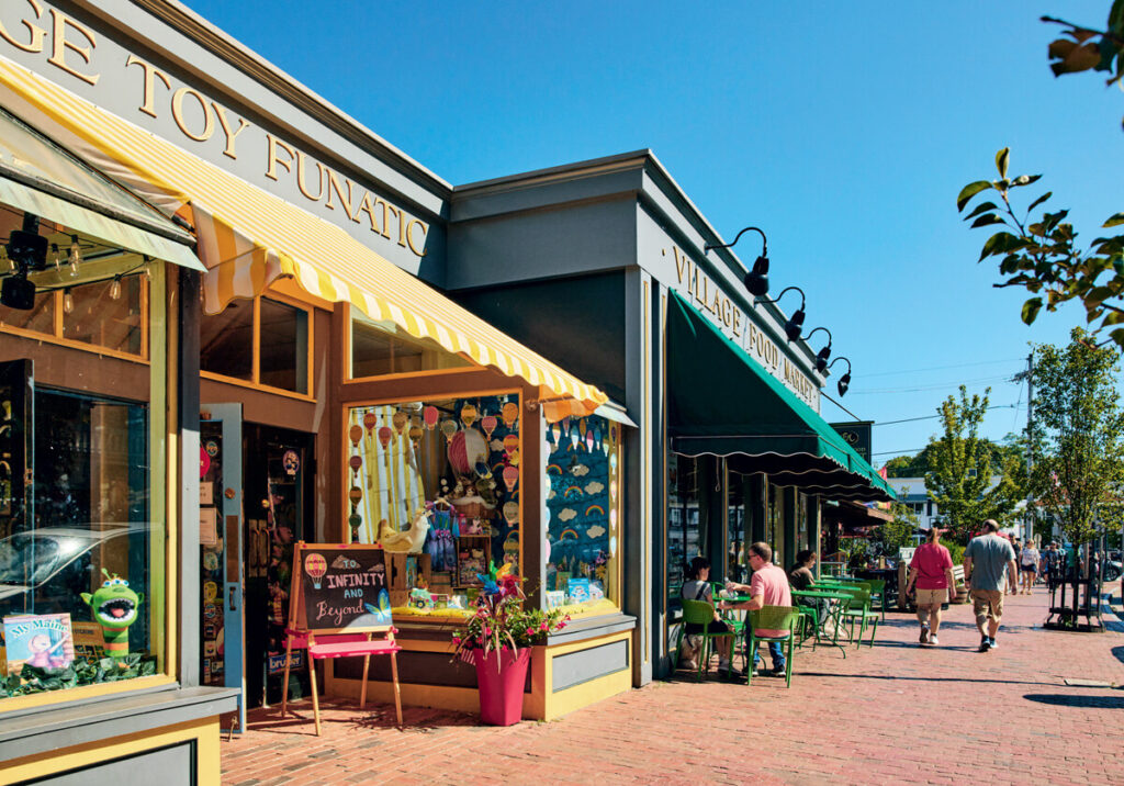 Colorful storefronts with outdoor seating and people walking on a sunny brick-paved street.