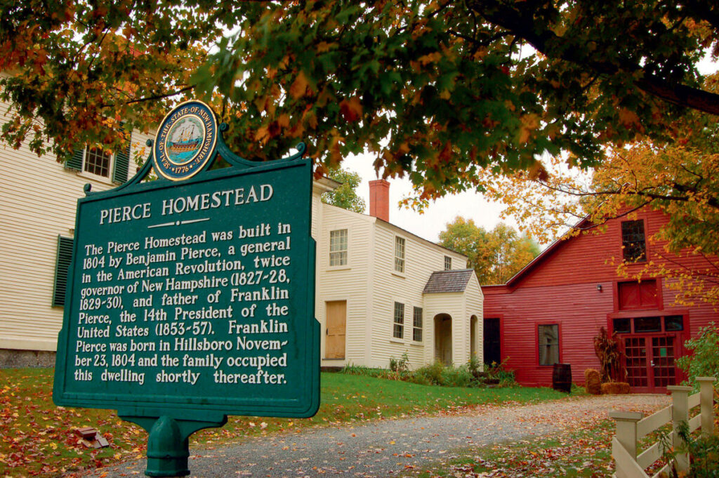 Historic sign for Pierce Homestead in front of two old homes with autumn trees in the background.