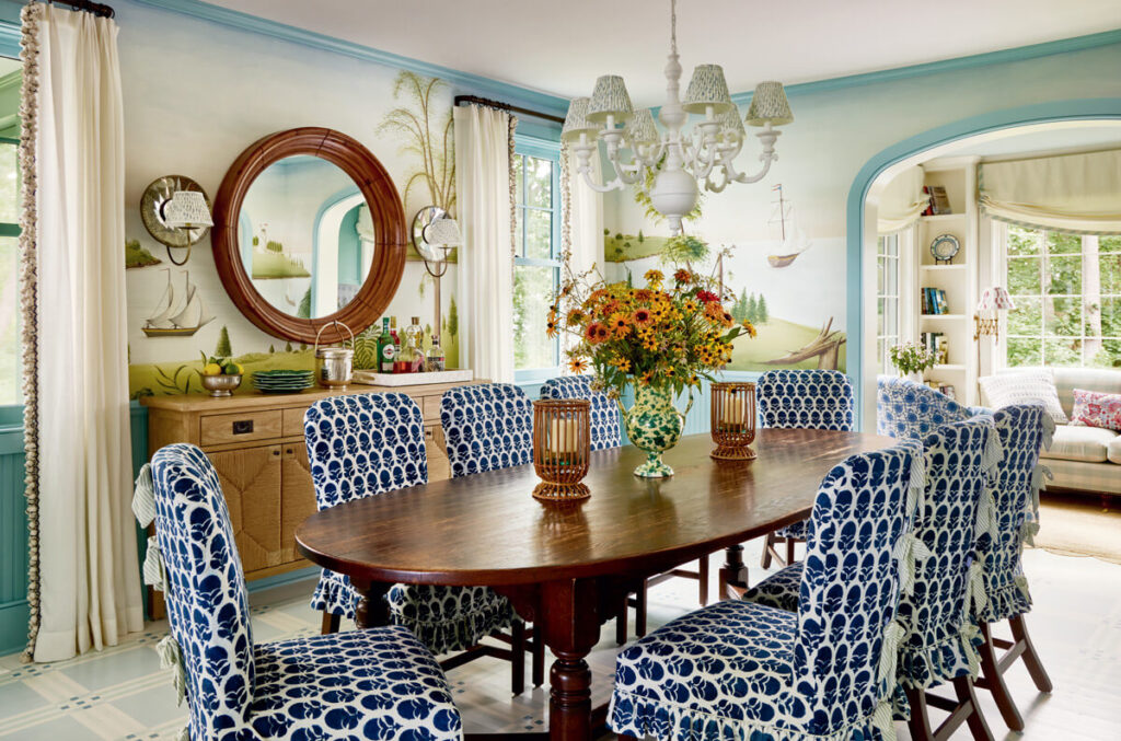 A dining room with a wooden table, blue-patterned chairs, flowers, and nautical-themed wall art.