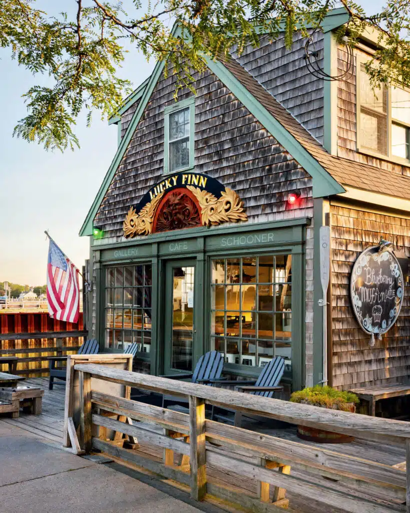 A rustic wooden shop called Lucky Finn sits by the water with benches, chairs, and an American flag outside.