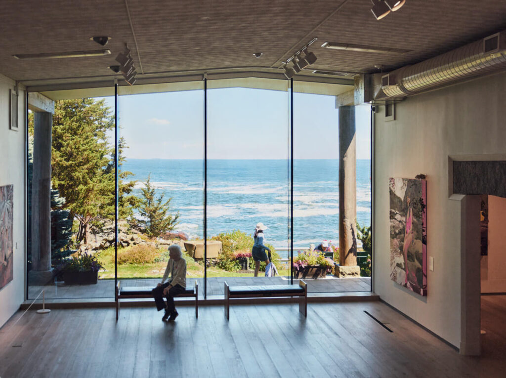 A person sits on a bench in a gallery with large windows overlooking the ocean and a garden outside.
