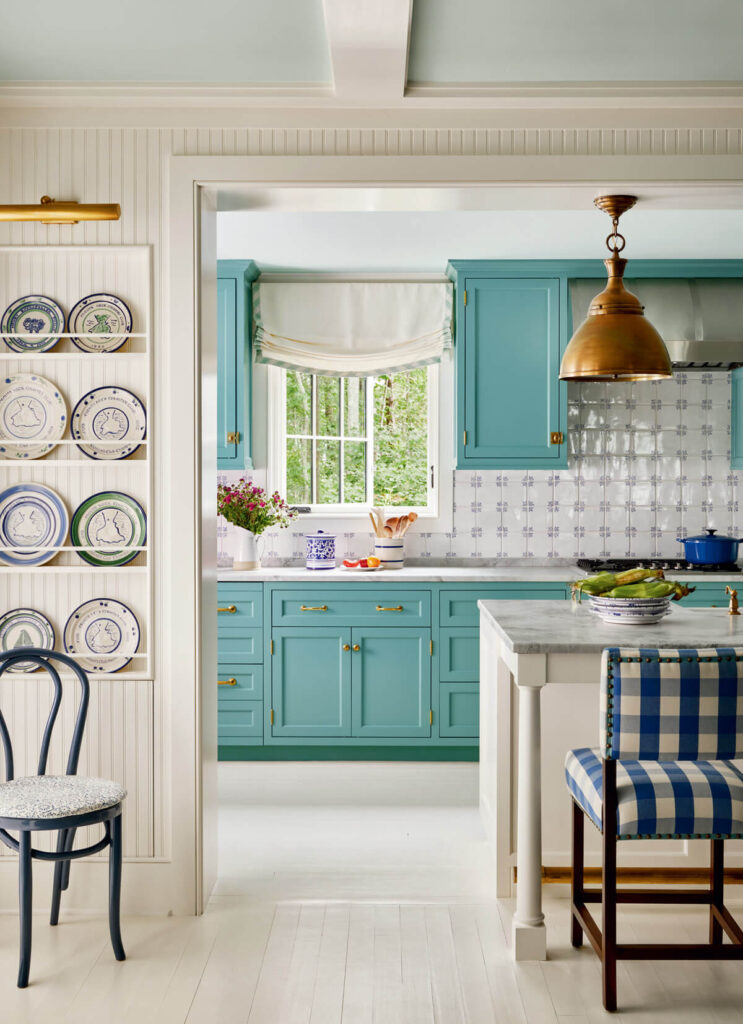 Bright kitchen with turquoise cabinets, gold hardware, patterned plates, and blue-check chair by a marble island.