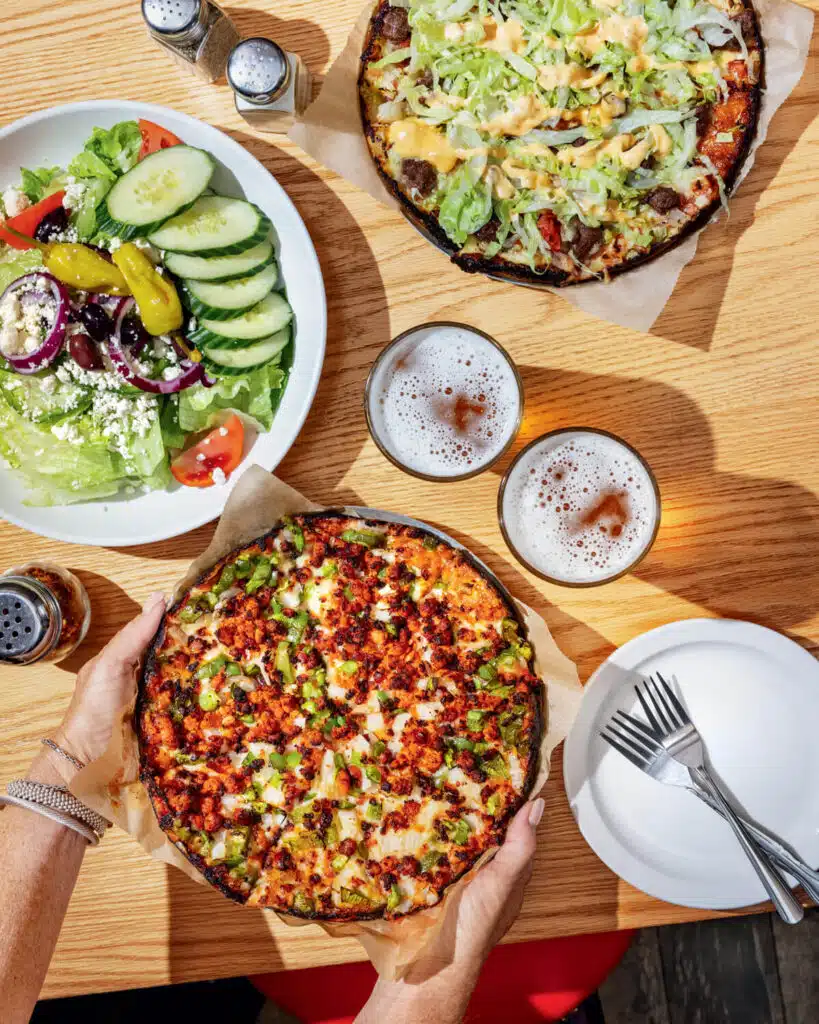 Two pizzas, a Greek salad, beers, and utensils on a wooden table, with hands holding one pizza.