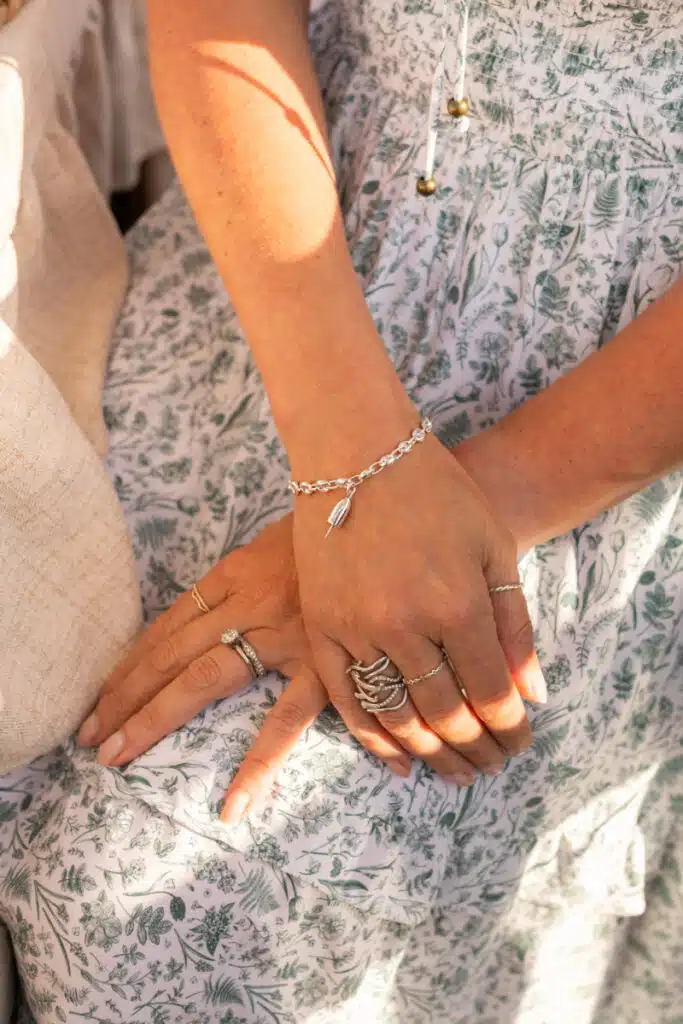 A woman with silver rings and a Patsy Kane bracelet rests her hands on her floral dress in sunlight.