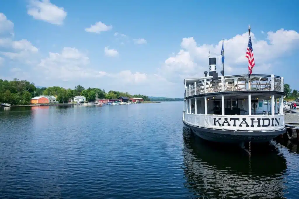 A white boat named Katahdin docked on a calm lake with buildings and trees in the background.