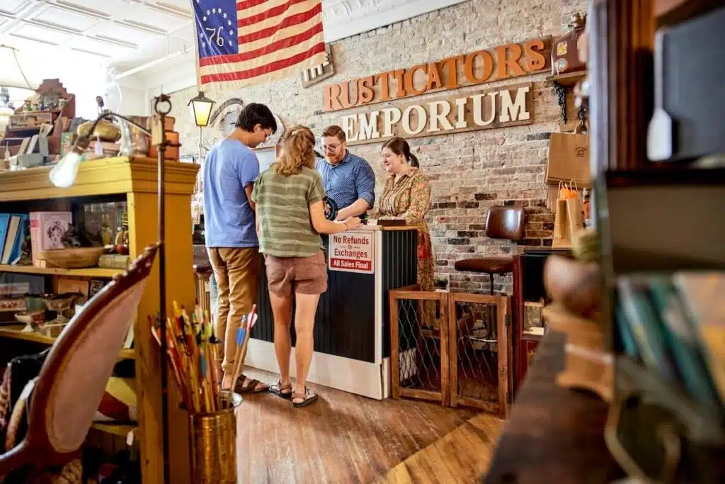 Two customers talk with two shopkeepers at a rustic emporium counter lined with various vintage items.