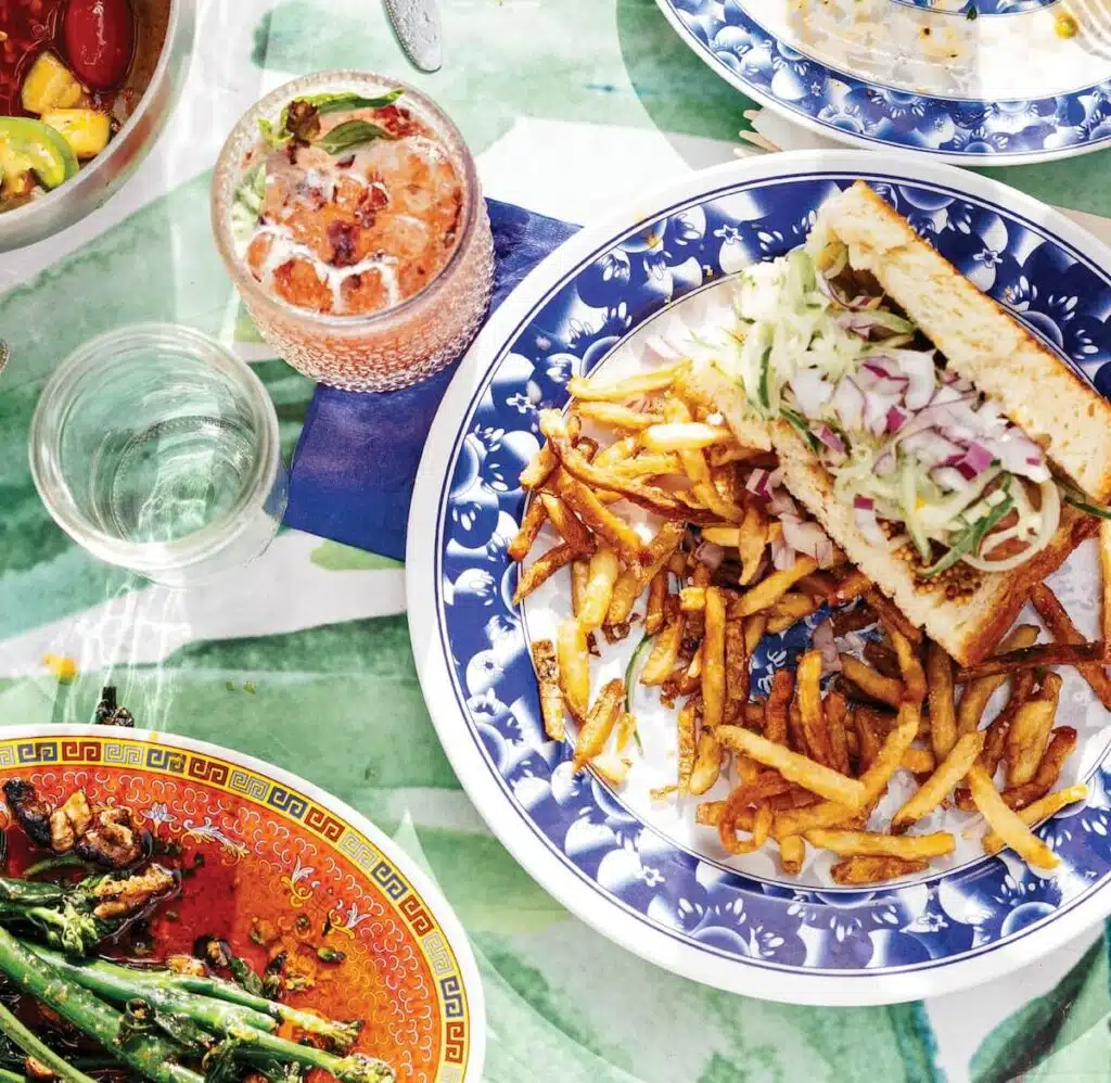 Sandwich with fries on blue plate, green beans in sauce, water glass, and pink drink on a green tablecloth.