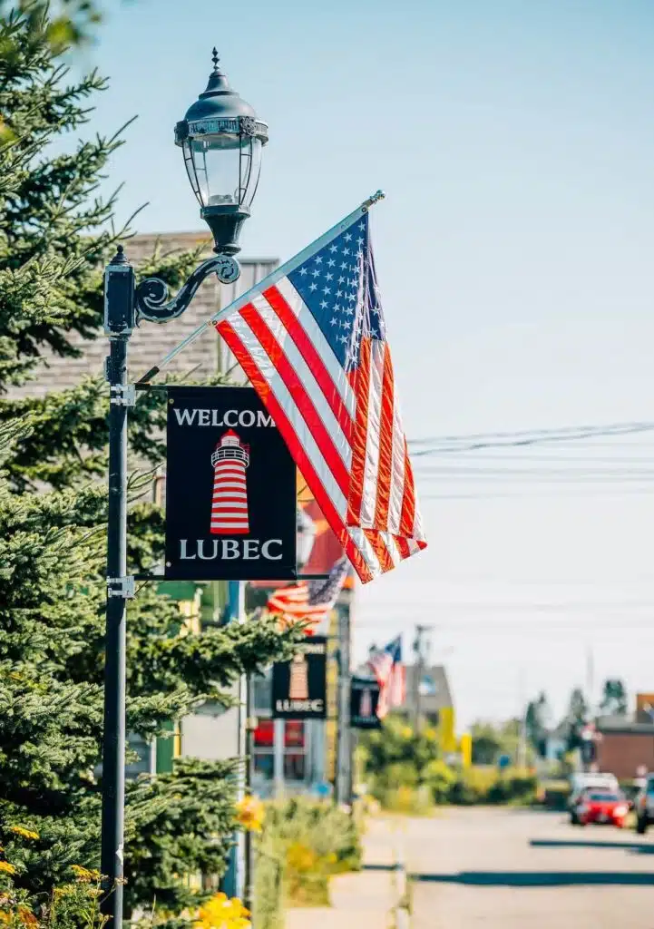 American flag and "Welcome Lubec" sign on a street with trees and lamp posts in the background.