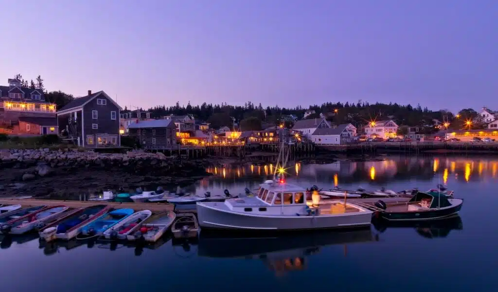 Harbor at dusk with boats docked, calm water, and warmly lit buildings along the shore.