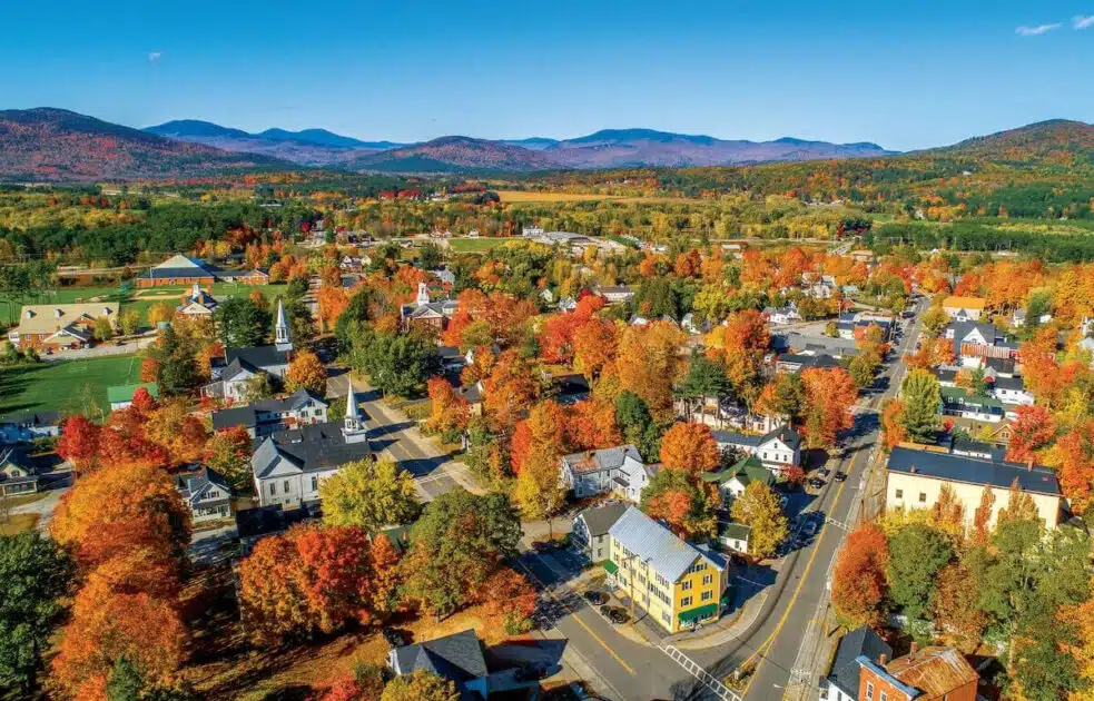 Aerial view of a small town with colorful autumn trees, houses, roads, and mountains in the background.