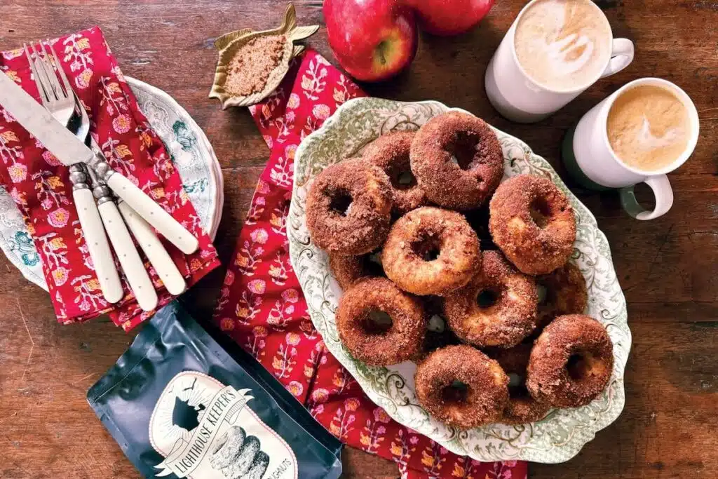 A plate of sugar-coated donuts, apples, coffee mugs, and napkins on a wooden table.