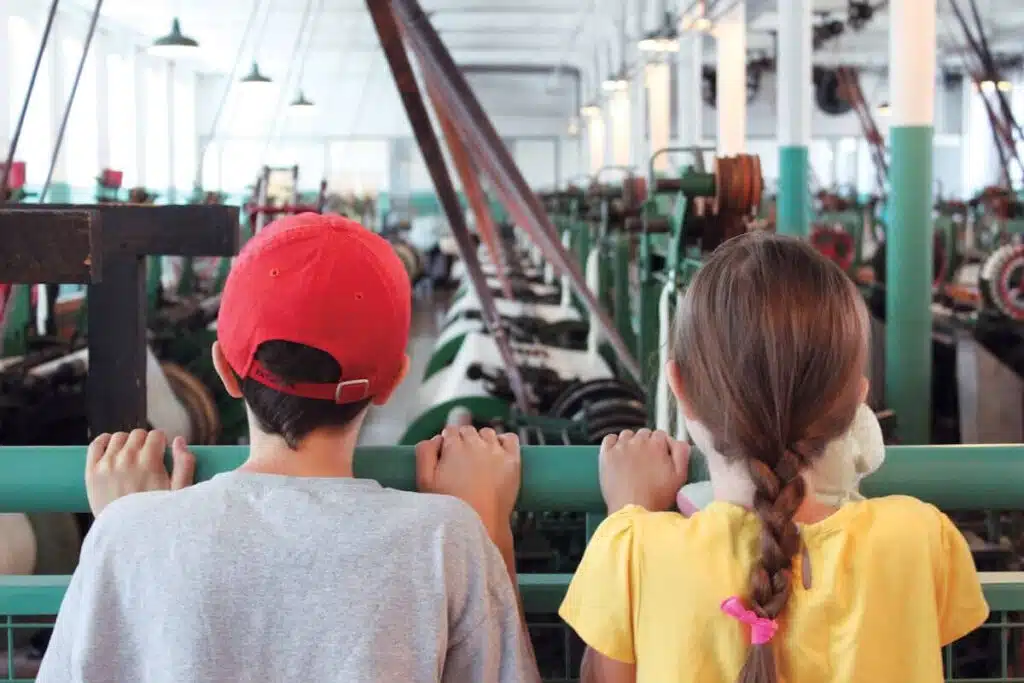 Two children look at machinery in a large industrial building, seen from behind.