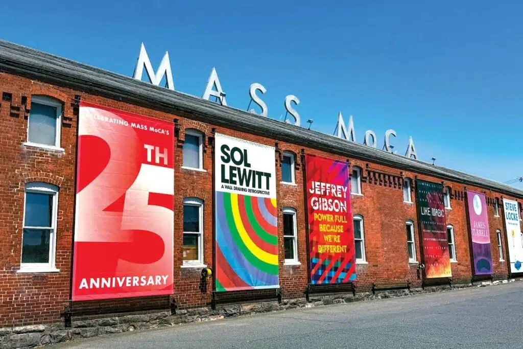 Large building with MASS MoCA sign and colorful banners for art exhibits on a sunny day.