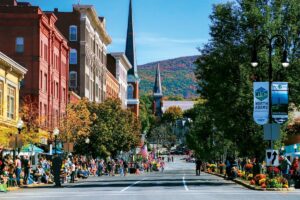 People gather along a tree-lined street with historic buildings and mountain views in North Adams, Massachusetts.