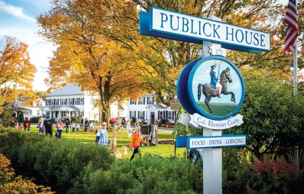 People gather on a lawn near the Publick House inn, surrounded by autumn trees and colonial buildings.