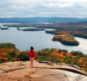 A child stands on a rocky cliff overlooking a lake with forested islands and autumn foliage.