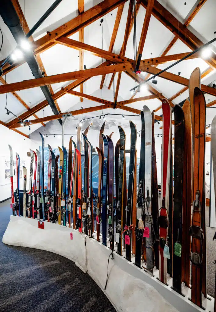 A display of vintage skis stands upright in a curved row inside a building with wooden ceiling beams.