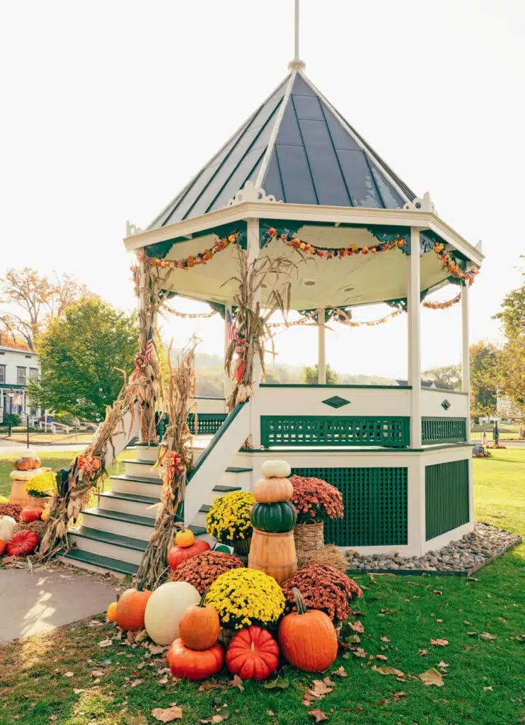 A gazebo decorated with autumn leaves, corn stalks, pumpkins, and mums on a sunny fall day.