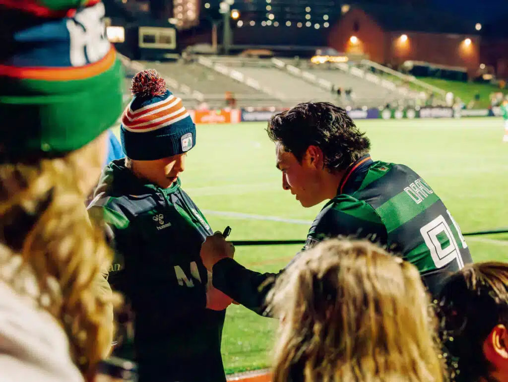 Rugby player in green signs an autograph for a fan at an outdoor stadium at night.