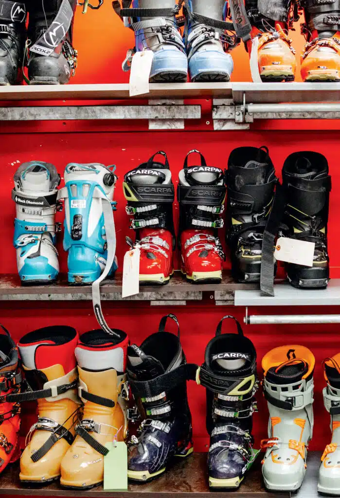 Rows of colorful ski boots on metal shelves against a red background, each pair with tags attached.