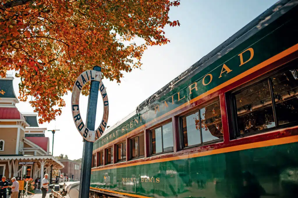 Vintage green train at a railroad crossing sign under autumn leaves, with people nearby and blue sky.