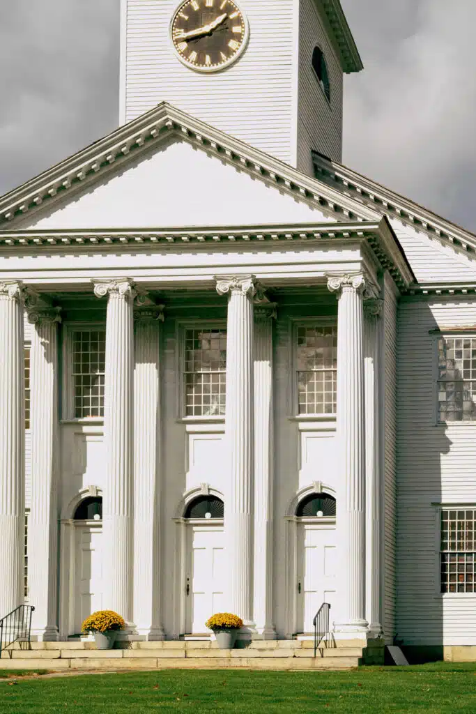 White building with tall columns, three doors, and a clock tower on a partly cloudy day.