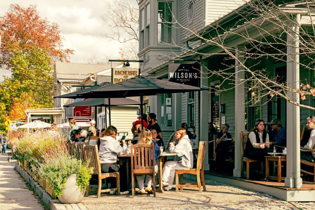 A Fall Escape to Connecticut’s Litchfield Hills. People sit at outdoor tables at a café on a sunny day; flowers line the sidewalk; trees in fall colors.