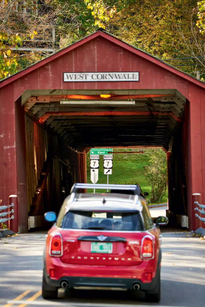 Guide to the Litchfield Hills. A red car enters the West Cornwall covered bridge with road signs visible in the background.