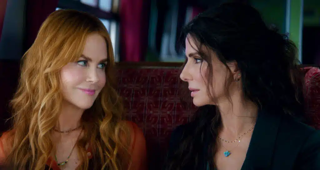 Two women with long hair sit closely on a red seat, looking at each other with soft expressions.