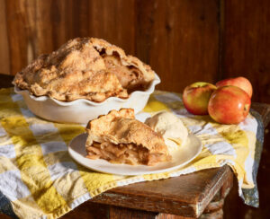 Blue Ribbon Apple Pie. A slice of apple pie with vanilla ice cream on a plate, with whole pie and apples in the background.