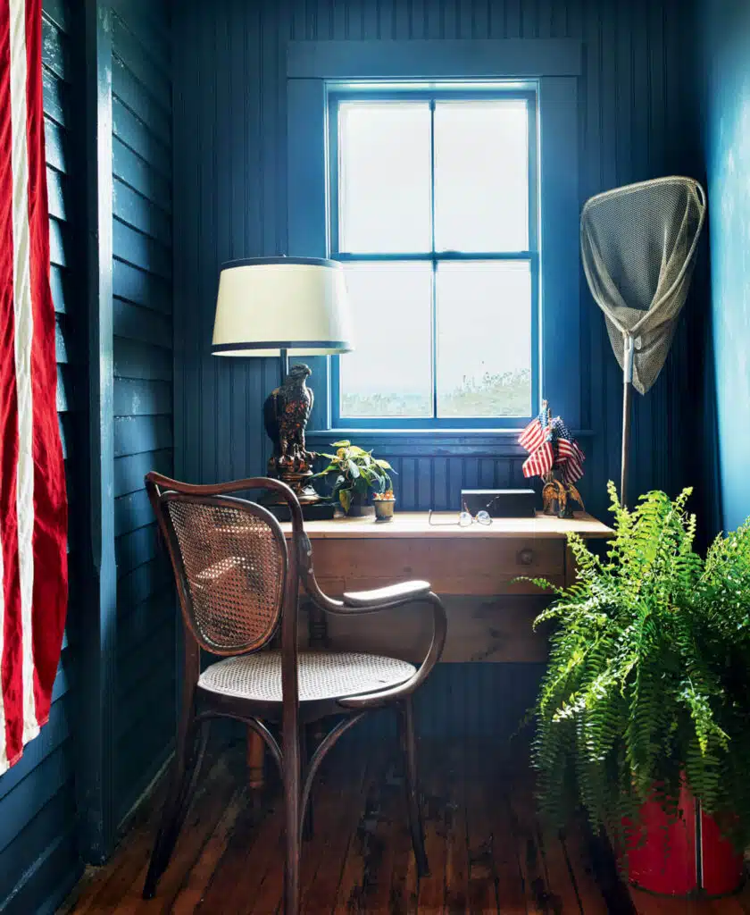 Small wooden desk with plants, a lamp, U.S. flags, and a chair by a bright window in a blue room.