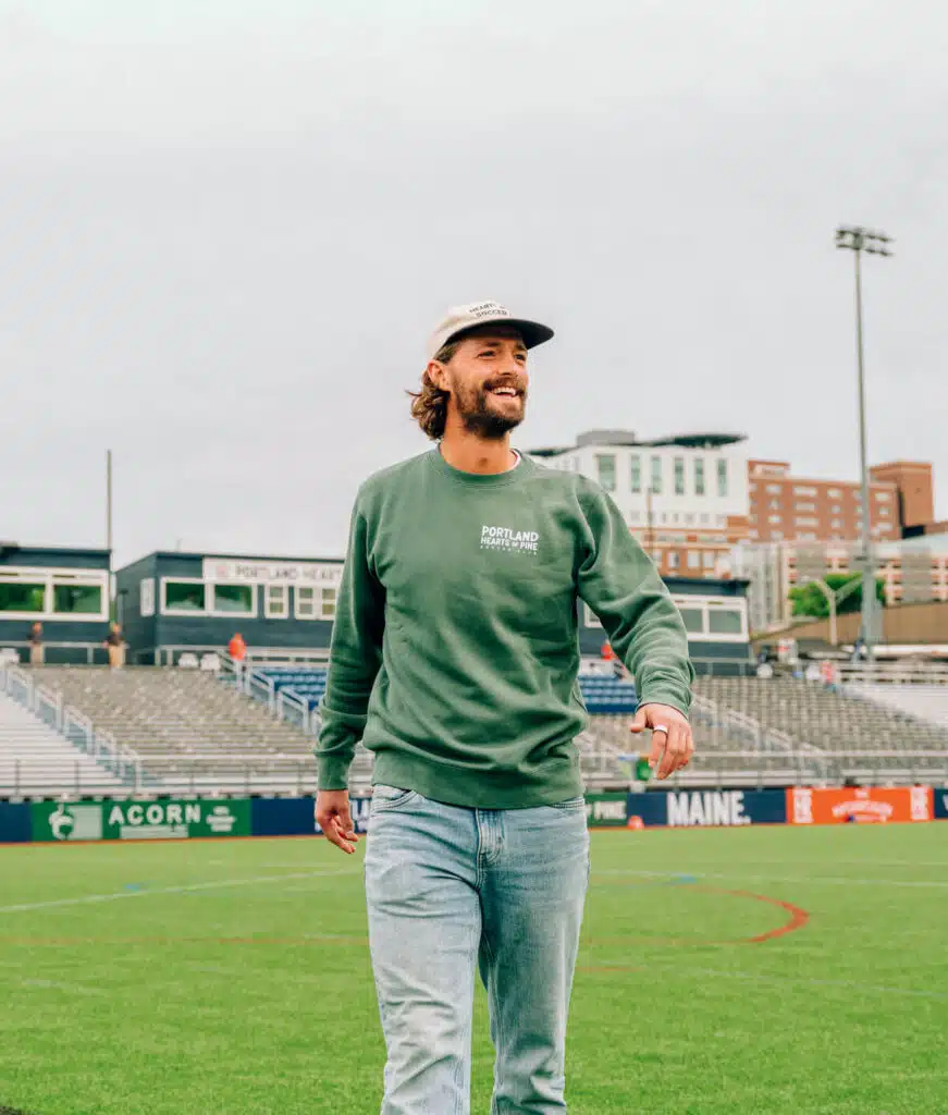 Hearts of Pine founder Gabe Hoffman-Johnson. Man in green sweatshirt and cap walking on sports field, stadium seats and buildings in the background.