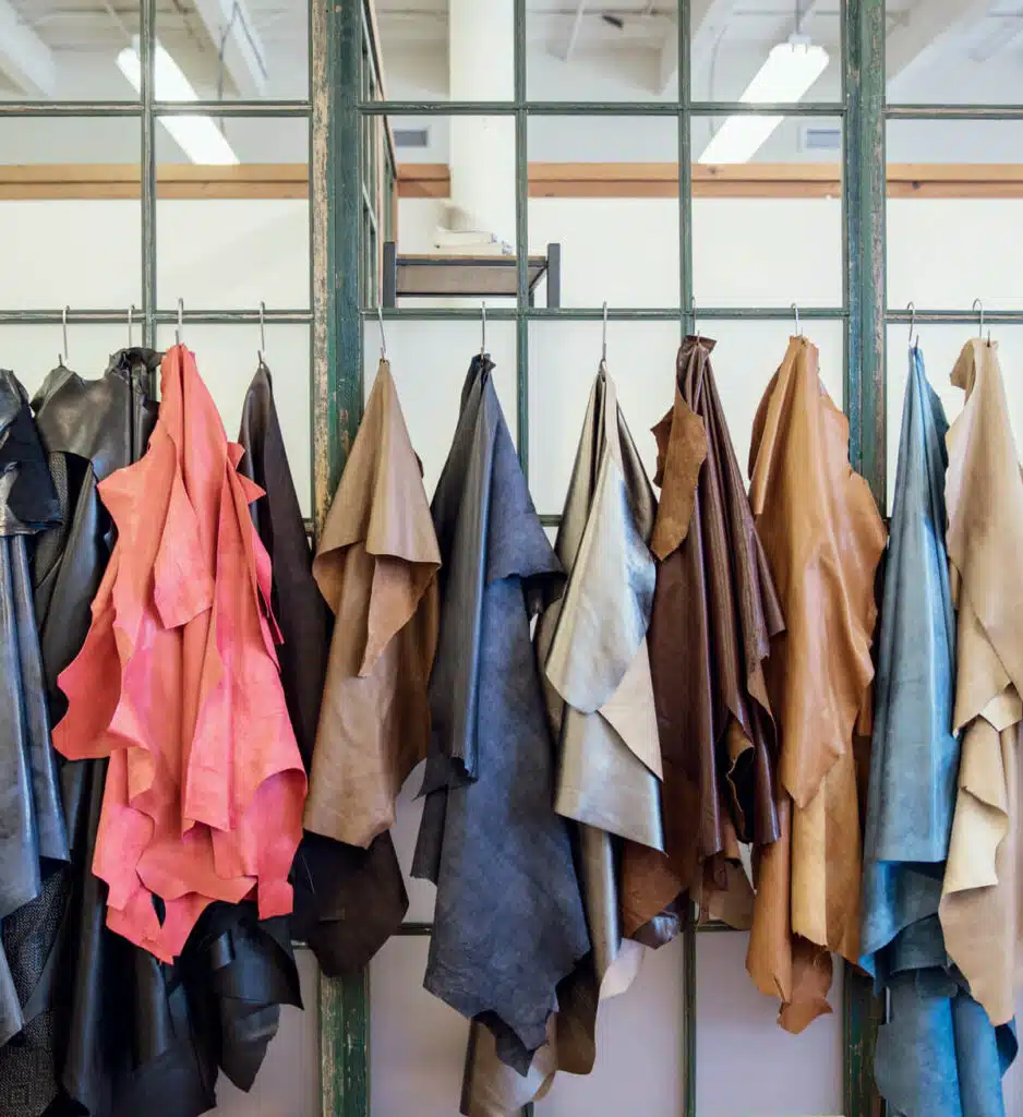 Various colored leather hides hang on hooks against a glass window with a metal frame.