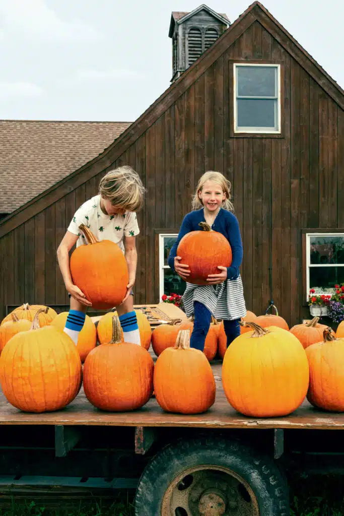 Two children hold pumpkins, standing on a wagon full of pumpkins in front of a brown wooden barn.