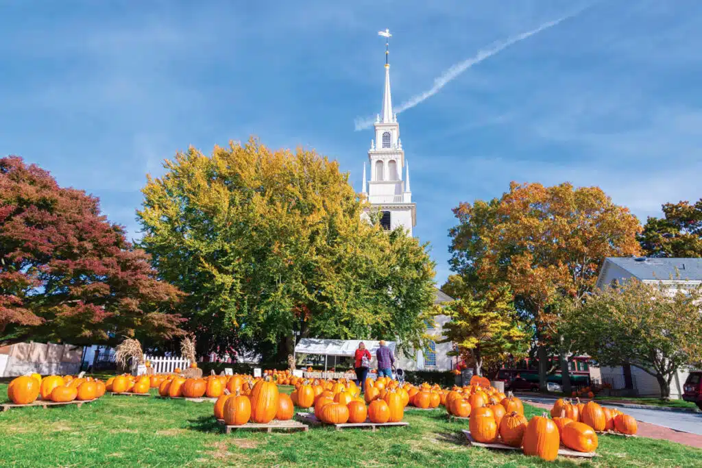 6 New England Foliage Adventures Worth the Drive This Fall. People walk among pumpkins on a grassy lawn with autumn trees and a white church steeple in the background.