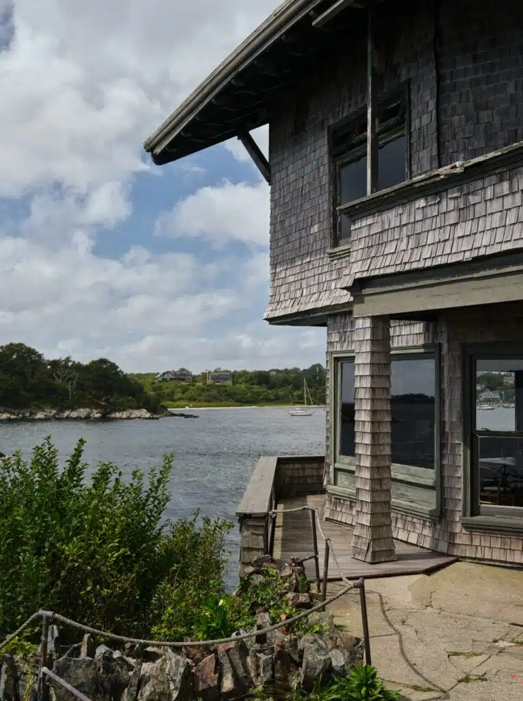 A weathered wooden house by a calm lake, with trees and a sailboat in the background under a partly cloudy sky.