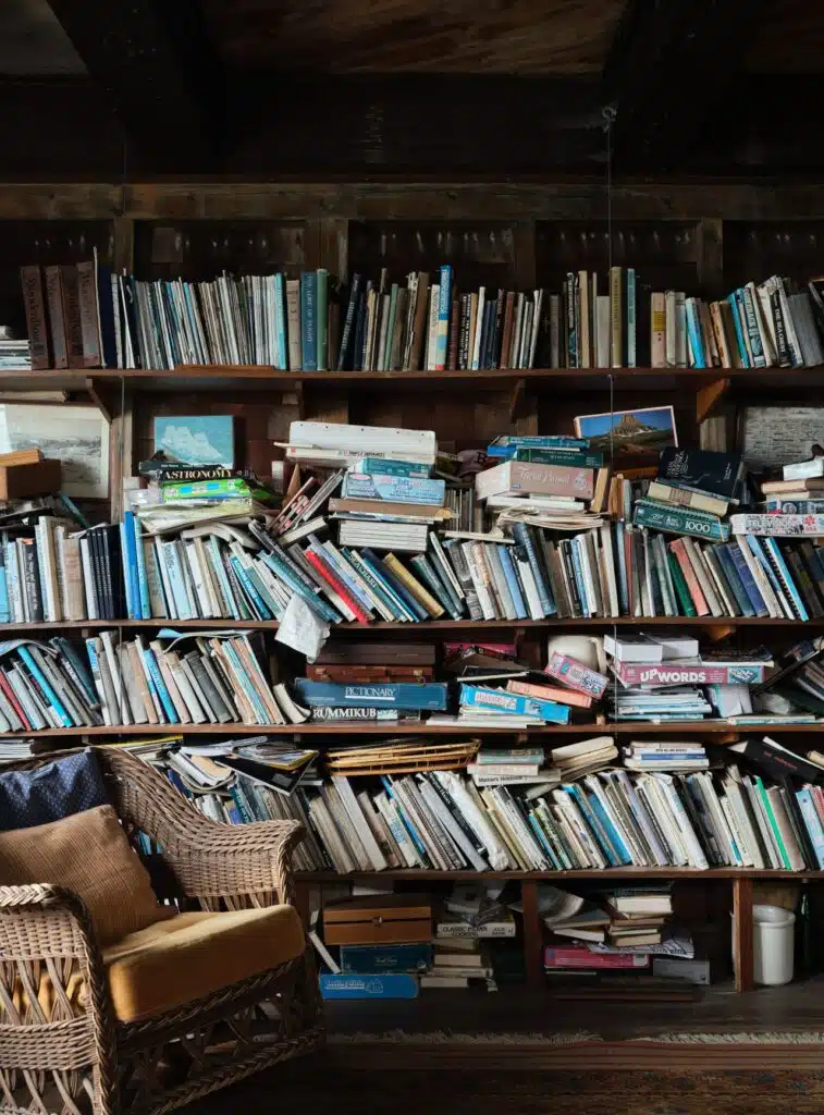 A wicker chair sits beside overflowing, disorganized bookshelves filled with assorted books and magazines.